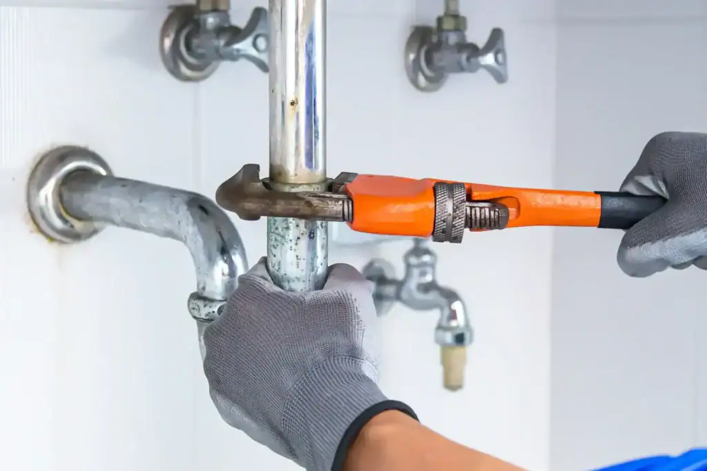 A plumber in Pasco County, FL, wearing gray gloves, uses an orange pipe wrench to tighten or loosen a metal pipe beneath a sink, with exposed plumbing and shut-off valves in the background.